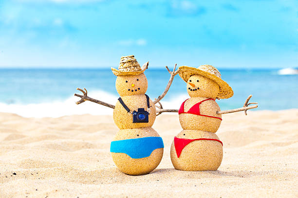 Sandman snowman couple winter vacationing on tropical beach of Kauai, Hawaii, USA. Tourists and honeymooners wearing swimsuit, straw hats, and camera, weaving happily, looking at the camera and smiling. Photographed in horizontal format with copy space on the right side of the photo.