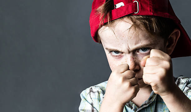 threatening 6-year old boy with freckles and a red hat back looking violent with fists in the forefront,acting like a little bully at school, contrast effects over grey background studio
