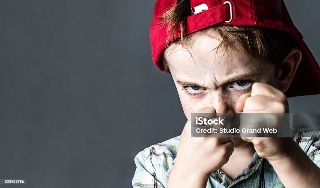 threatening boy with freckles and red hat back looking violent threatening 6-year old boy with freckles and a red hat back looking violent with fists in the forefront,acting like a little bully at school, contrast effects over grey background studio Child Stock Photo