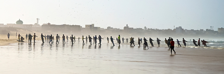 A row of Senegalese fishermen are pulling in their nets on the beach. There is a sense of teamwork and togetherness, on the side there are people exercising. On the horizon is the silhouette of Dakar and a mosque on the left side. There is a haze and the men are silhouettes.