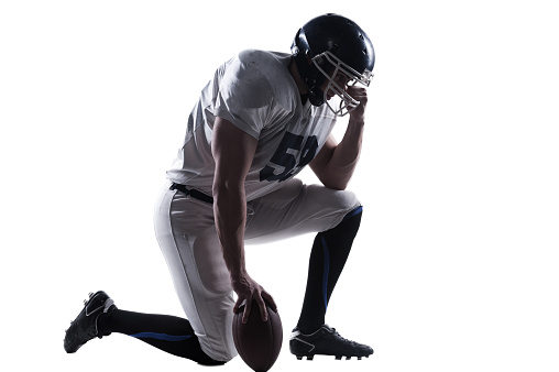 Side view of American football player holding hand on helmet while standing on knee against white background
