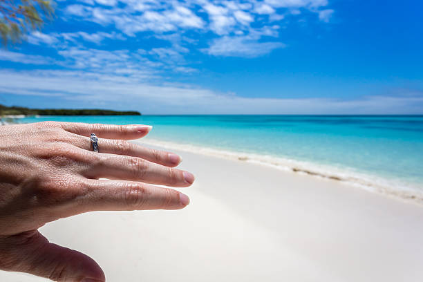 Woman hand with engagement ring on tropical beach. The water is clear and the sky is clear with few clouds. 