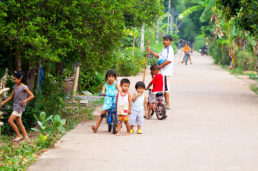 Parangmee, Thailand - May 29, 2013: Cature of some little Thai kids playing on road in a very  small village in countryside of Phitsanulok. A girl is sitting on bicycle. A boy in background is also on bocycle. A man is standing behind children.