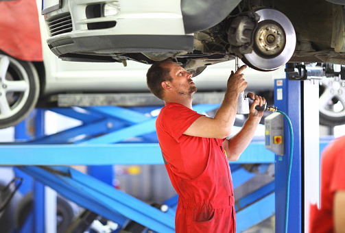 Adult male car mechanic replacing break system on vehicle in car service facilities. Row of cars behind him, blurry. He's wearing red working uniform.