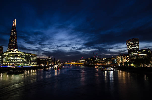 Night view of Thames River from London Tower Bridge. On left we see the Shard building, and on the right the Walkie Talkie building. The sky is a dark blue and buildings are spreading their lights into the river.