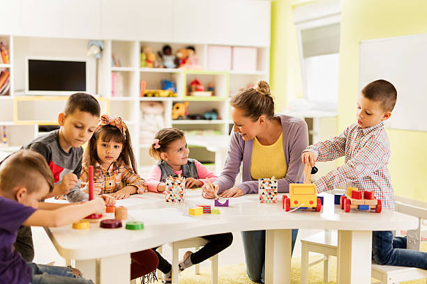 Happy teacher enjoying with group of children at preschool. Happy teacher sketching and talking to a smiling little girl at kindergarten while other children are playing with educational  toys. preschool building stock pictures, royalty-free photos & images