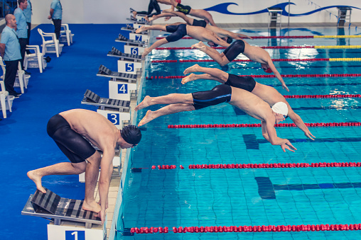 Group of swimmers in mid-air jumping into the pool while one is left behind still crouching on the starting block because of bad reflexes.