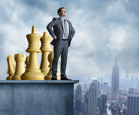A very confident businessman standing on top of a building with his team of chess pieces behind him.