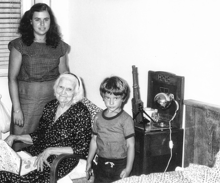 Grandmother and nephews inside bedroom in 1970. The Grandmother is sitting on armchair and the nephews are standing close to her. Some skratches and grain due to the age of the photo. Scanned black and white print. 