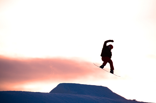 A teenaged male snowboarder hits a jump at sunset while riding at a winter ski resort.