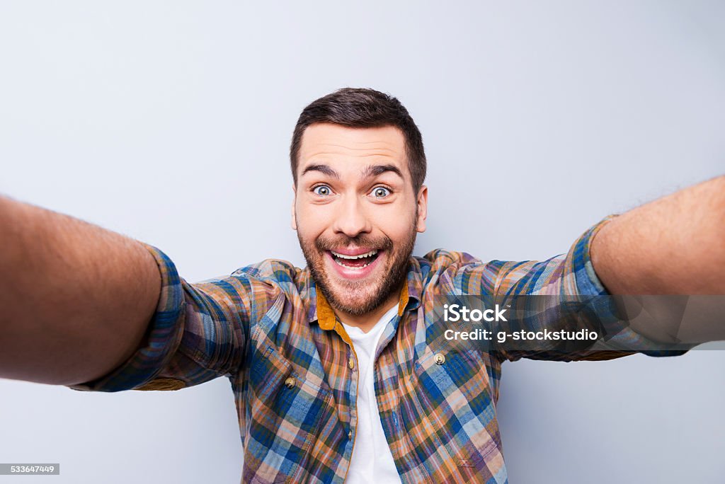 I love selfie! Handsome young man in shirt holding camera and making selfie and smiling while standing against grey background Selfie Stock Photo