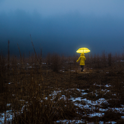 Little girl in the yellow coat with umbrella crossing the field in fog. Homage to the Little Red Riding Hood