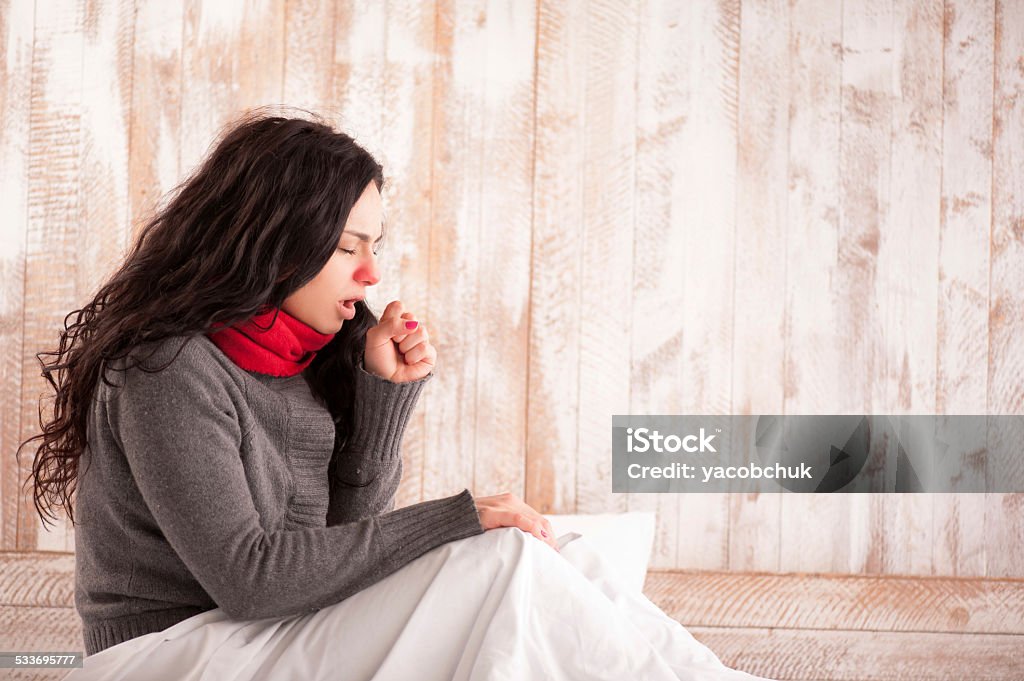 Young beauty caught a cold Coughing beauty. Side view image of young sick woman with scarf on her neck sitting in bed and coughing with her country house on the background Coughing Stock Photo