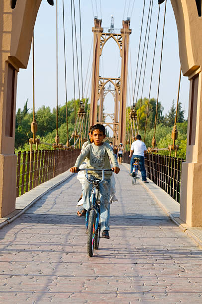 Suspension bridge in Deir ez-Zur, Syria Deir ez-Zur, Syria - June 20, 2010: Two Syrian boys ride a bicycle over a suspension bridge built above the Euphrates River. The bridge was built by the French in 1927, and would be destroyed by shelling during the Syrian conflict in 2013. Other people are visible in the background crossing the bridge. euphrates syria stock pictures, royalty-free photos & images