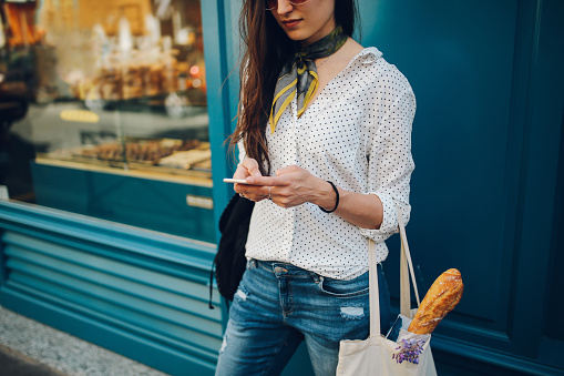 Vintage toned image of a young Parisian woman using her smartphone while walking the streets on Montmartre district of Paris. She is wearing a white dotted pattern shirt and sunglasses, carrying a canvas tote bag after some shopping for groceries and food. Paris, lifestyle, street style fashion concepts.