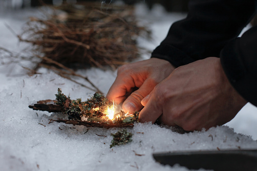 Man lighting a fire in a dark winter forest, preparing for an overnight sleep in nature, warming himself with DIY fire. Adventure, scouting, survival concept.