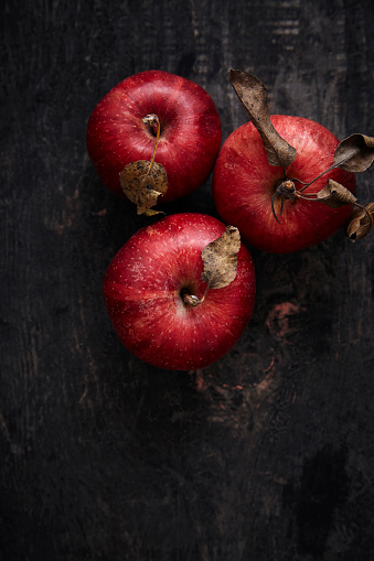 Overhead view of old dark wooden table and organic red apples on the middle.This an old rustic table.Weathered and worned dark piece of cut wood. There are scratches and stains on wood.There are three vivid colored apples standing together.This a daylight shot image and focus is on apples.