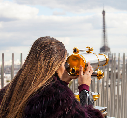 Rear view of young woman standing on Arc de triomphe and using a telescope to see the Eiffel tower. Wears fur coat. Looking at her reflection on the telescope. Telescope in gold and silver. Eiffel tower and a railing on background. Cloudy sky.