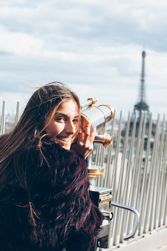 Young woman standing on Arc de triomphe and using a telescope to see the Eiffel tower. Wears fur coat. Looking at camera. Telescope in gold and silver. Eiffel tower and a railing on background. Cloudy sky.