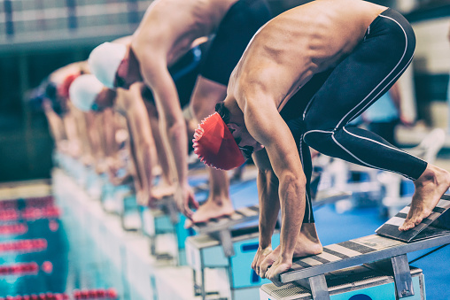 Close up shot of a male swimmer crouching on starting blocks ready to jump into the water at the signal, other swimmers defocused.