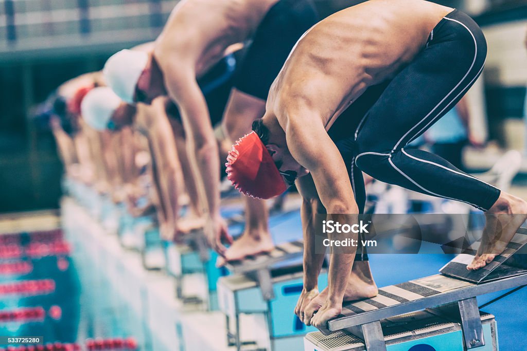 Swimmer crouching on starting block ready to jump Close up shot of a male swimmer crouching on starting blocks ready to jump into the water at the signal, other swimmers defocused. Swimming Stock Photo