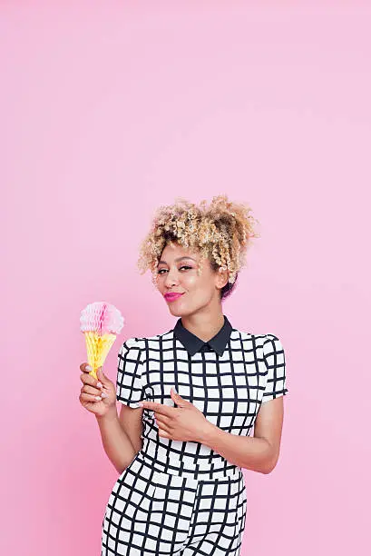 Summer portrait of happy, afro american young woman wearing grid check playsuit, standing against pink background, holding ice cream honeycomb decorations and smiling.