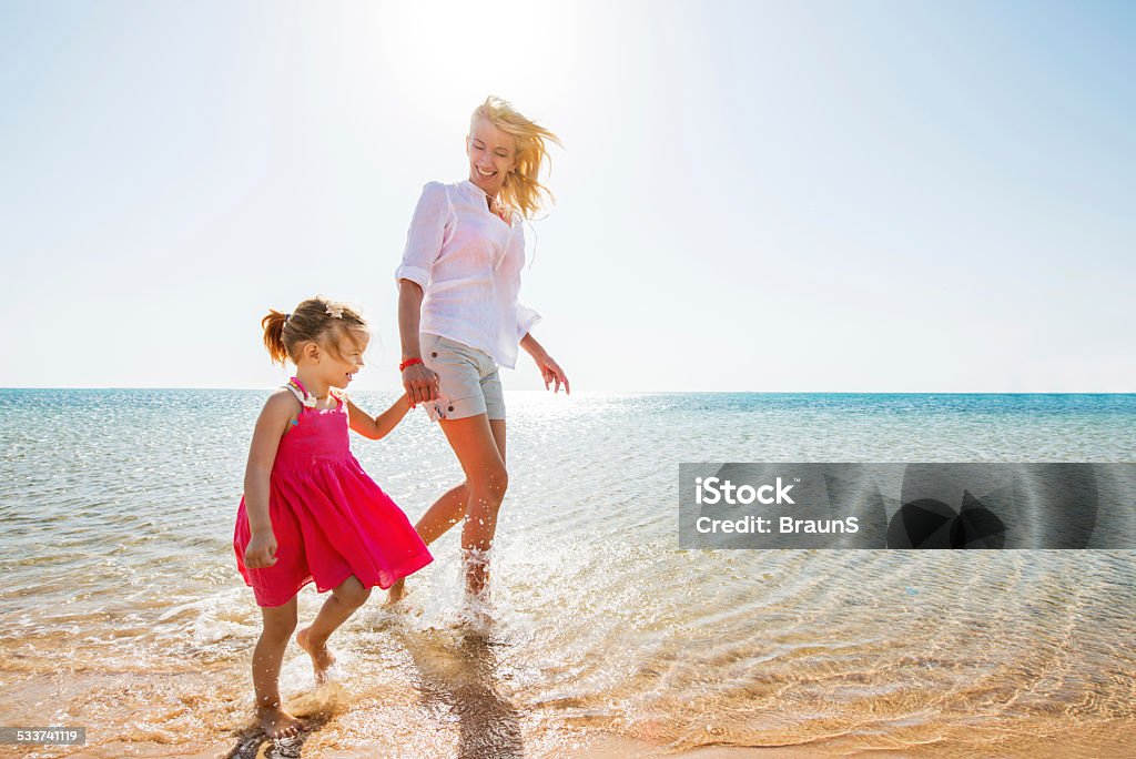 Mother and daughter walking at the beach. Happy mid adult mother enjoying in summer day with her little girl. They are holding hands and walking through water. Child Stock Photo