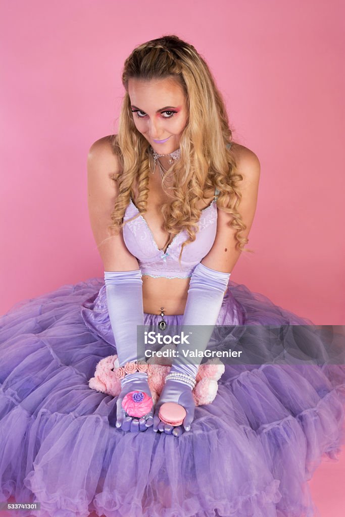 Young woman sitting on floor offering sweet treats. Vertical studio shot on pink of young blonde woman in purple crinoline, bra and gloves, offering a cupcake in one hand and a pink macaroon in the other. 20-29 Years Stock Photo