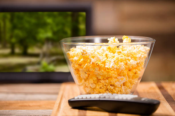 Watching show, movie on television at home. Popcorn, remote control. Watching a show or movie on television at home. Popcorn in clear bowl with TV remote control on coffee table. Forest scene on TV screen background. remote control on table stock pictures, royalty-free photos & images