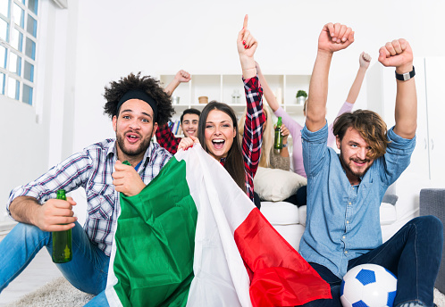 Group of young people sitting on a sofa watching a sporting event on TV and cheering. One of the men is holding a ball and two of the them are holding glass bottles. Young woman holding Italian flag