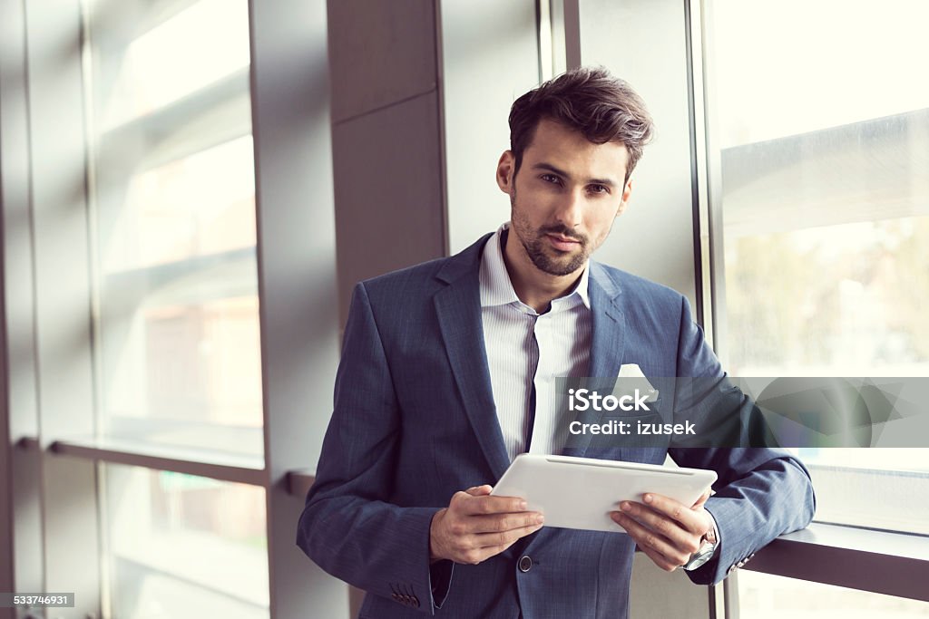 Elegant businessman holding a digital tablet Businessman wearing suit standing by the window in an office, holding a digital tablet in hands and looking at camera. 2015 Stock Photo