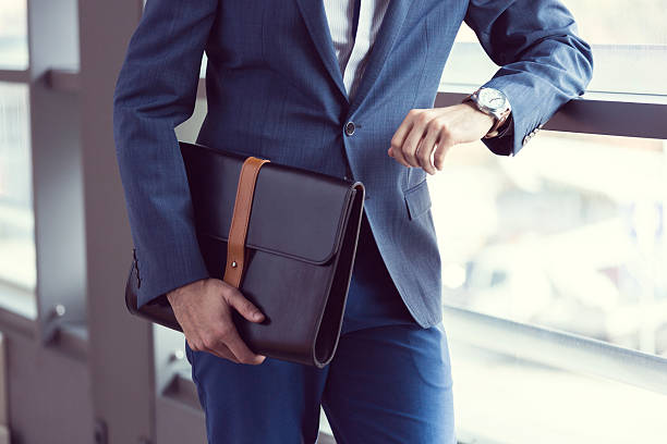 Elegant businessman in the office Businessman wearing suit posing in an office, holding leather briefcase and checking the time. briefcase stock pictures, royalty-free photos & images