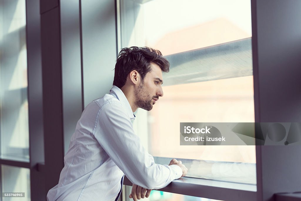 Young businessman looking out the window Young businessman wearing white shirt looking out the window in an office. Businessman Stock Photo