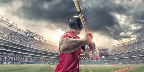 A close up rear view of a professional male baseball player wearing a red baseball strip and safety helmet, holding baseball bat just about to strike incoming baseball. The action occurs during a baseball game in a generic floodlit outdoor stadium full of spectators. Baseball ground, scoreboard etc are all fictional.