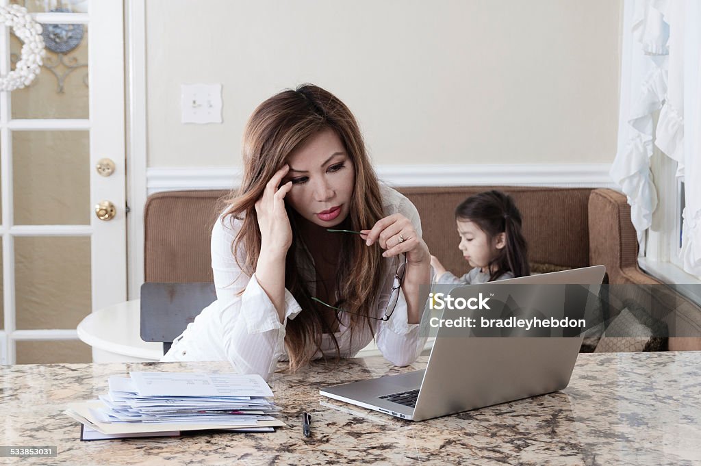 Worried single mother under financial strain An attractive Asian mother works from home, specifically from her kitchen. Her five year old daughter can be seen behind her, eating at the kitchen table. Her Macbook lays on the granite kitchen counter, along with a stack of papers and documents. She appears distressed/worried about something as she looks at her laptop screen.  Distraught Stock Photo