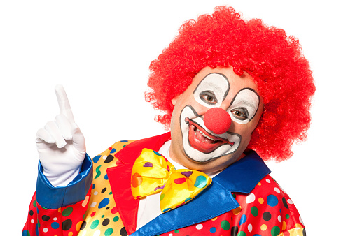 Portrait of a funny playful female clown in colorful wig holding lollipops in both hands, looking at camera and smiling, isolated on a white background