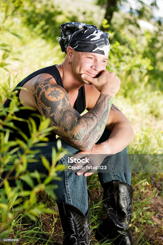 Characters: Biker man with tattoos and piercings contemplates. Nature. Tribal man or biker with heavy tattoos on his arm and several piercings contemplates, meditates, smiling. Nature, Outdoors, Forest. He wears a head scarf and cowboy boots.   2015 Stock Photo