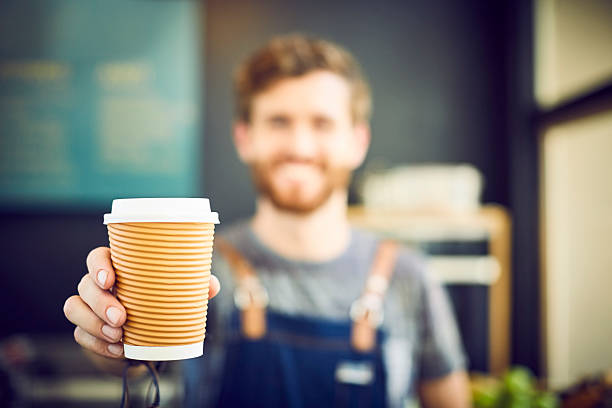 Young barista offering coffee in disposable cup. Smiling waiter is serving drink in coffee shop. Focus is on container.