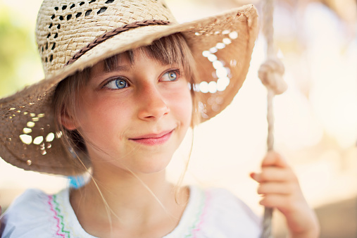 Cute little girl having fun on a swing in summer. The girl is wearing cowboy hat and is smiling delicately.
