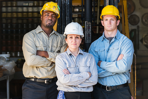 A multiracial group of three workers in their 30s, standing in a factory with their arms folded, looking at the camera.  They are wearing hard hats, standing in front of a forklift, with various tools and supplies in the background.  The foreman, standing in the middle, is a woman, looking serious and confident.