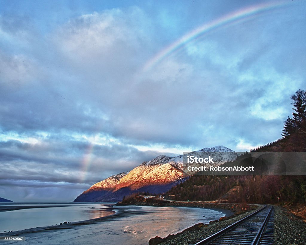 Turnagain Arm Sunrise Train tracks, snow covered mountains and the Seward Highway are illuminated by the sunrise and a rainbow over Turnagain Arm in south central Alaska. Snow Stock Photo