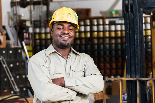 A happy African American man working in a printing factory.  He is standing with arms crossed in the storage warehouse of the small business, next to a forklift.