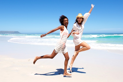 Full body portrait of two young women friends laughing and running on the beach