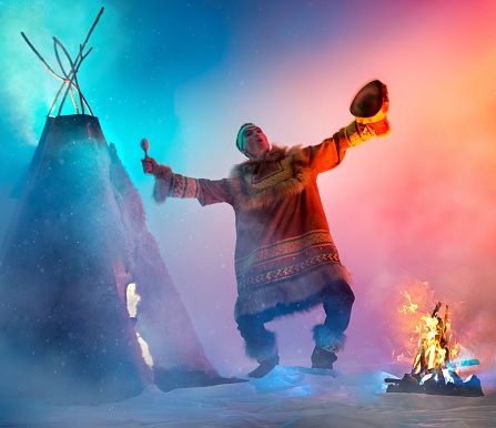The young man with appearance of Siberian indigenous people next to a bonfire. The attractive man dressed ethnic clothing. He singing, dancing folk dance and drumming in shaman drum. Dance performance in ethnic decoration. Studio shooting