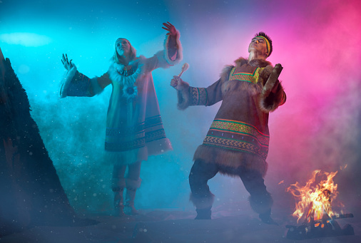 The beautiful young woman and young man with appearance of Siberian indigenous people are dressed in ethnic clothing.  The blonde young woman and attractive man are dancing a folk dance. The man is drumming in shaman drum. Theatrical performance in ethnic decoration, studio shooting in smoke