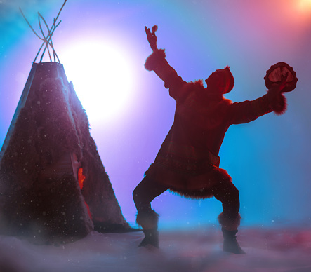 The silhouette of young man on backdrop of a aurora borealis. The attractive man dressed ethnic clothing of Siberian indigenous people. He singing, dancing folk dance and drumming in shaman drum. Dance performance in ethnic decoration. Studio shooting