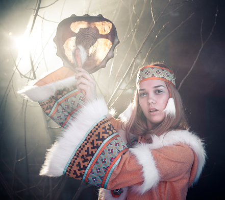 The beautiful young dressed ethnic clothing of Siberian indigenous people is dancing folk dance and drumming in shaman drum. The blonde young woman is holding drum high looking at the camera. Theatrical performance, studio shooting in smoke on black background