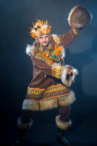 The mature man with appearance of Siberian indigenous people. The serious man with mustache dressed ethnic clothing. He is dancing folk dance and drumming in shaman drum. Studio shooting in smoke on black background