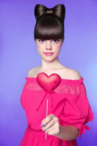 Attractive teen girl with bow hair style, brunette young model holding red heart isolated in blue background. studio portrait