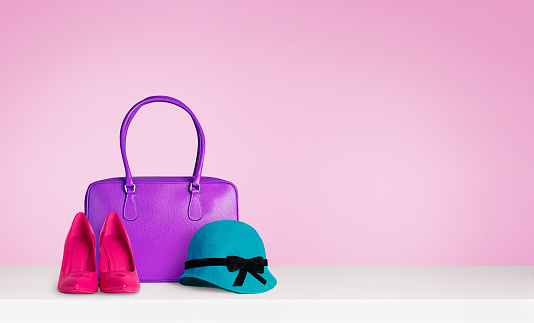 Purple leather handbag, red pink high heels, and green hat on white table.